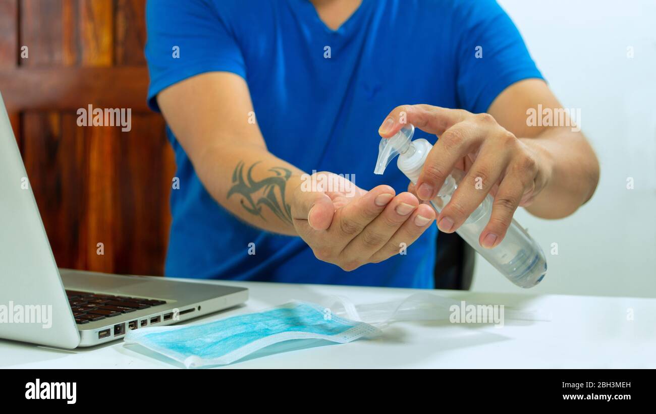 Close up on a man in a blue shirt sitting washing his hands with a plastic bottle of alcohol gel, working with his laptop and a blue mask on the table Stock Photo