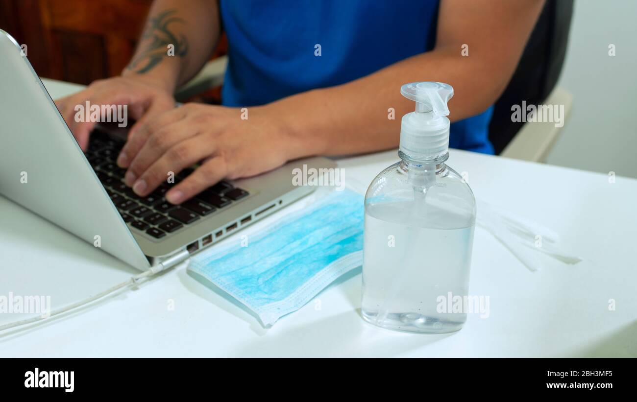 Close up to a plastic bottle of alcohol gel next to a blue face mask on a white table next to a man in a blue shirt sitting working with his laptop Stock Photo