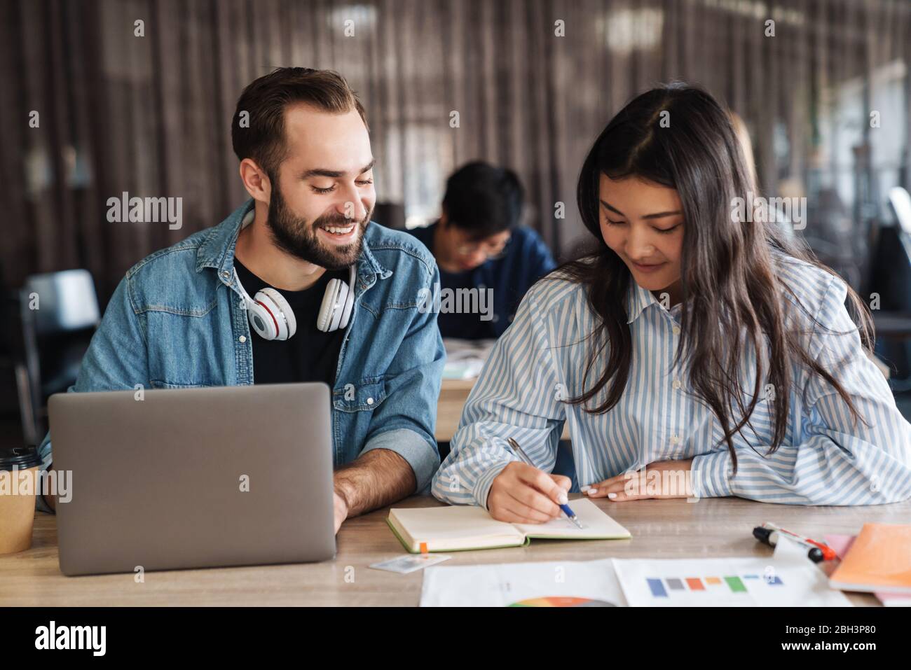 Photo Of Multinational Joyful Students Using Laptop And Writing In Exercise Book While Studying At Classroom Stock Photo Alamy