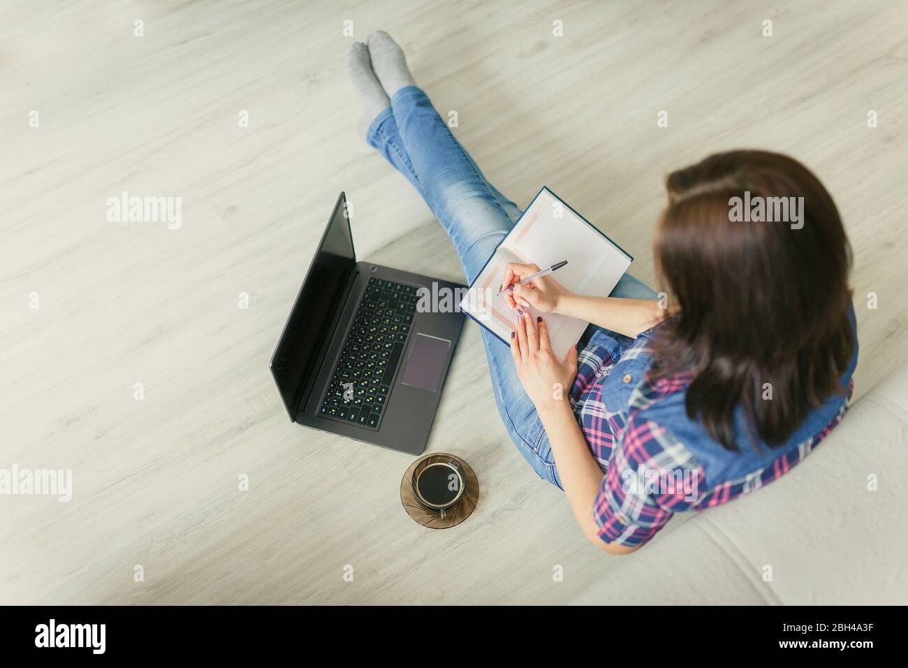 The girl sitting at home on the floor with a laptop, a cup of coffee and making diary entries.Homeworking, freelance. Social distance concept.Quaranti Stock Photo