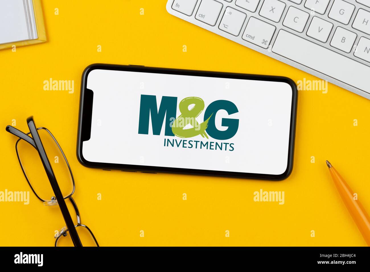 A smartphone showing the M&G Investments logo rests on a yellow background along with a keyboard, glasses, pen and book (Editorial use only). Stock Photo