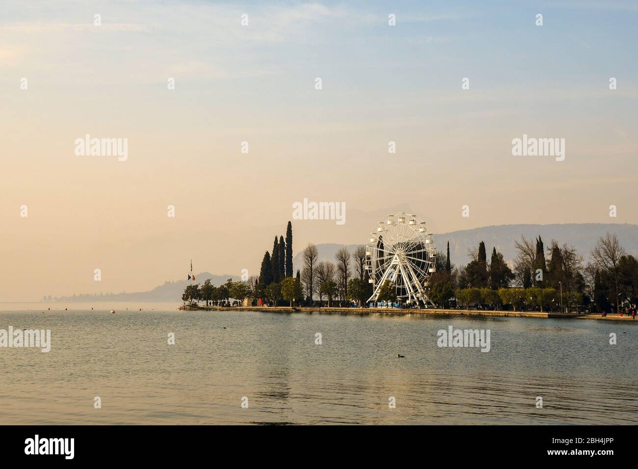 Scenic View Of Lake Garda With A White Panoramic Wheel On The Lakefront scenic-view-of-lake-garda-with-a-white-panoramic-wheel-on-the-lakefront
