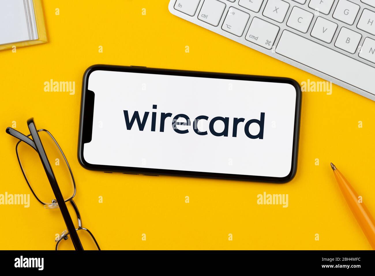 A smartphone showing the Wirecard logo rests on a yellow background along with a keyboard, glasses, pen and book (Editorial use only). Stock Photo