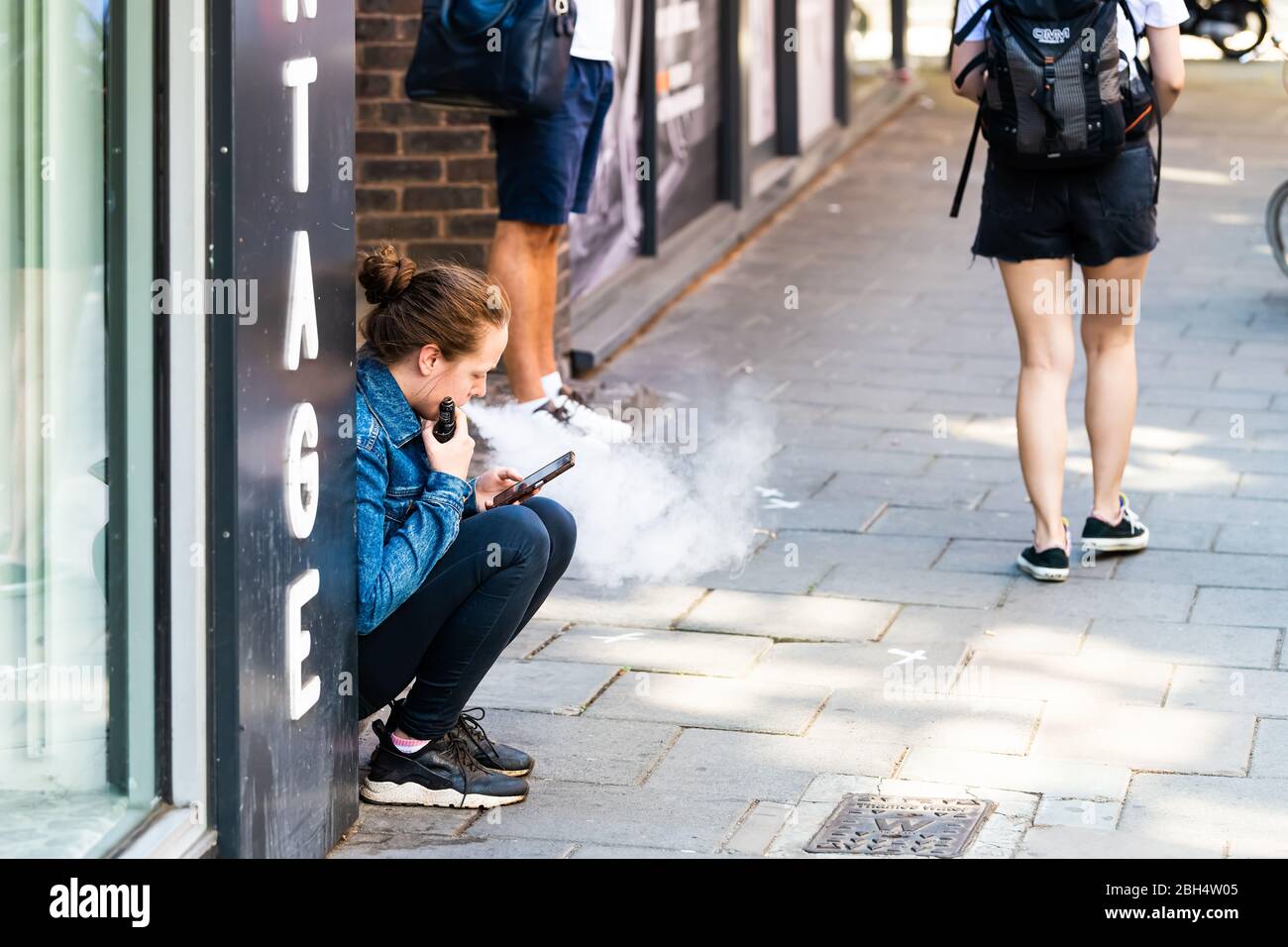 London, UK - June 26, 2018: People woman sitting on sidewalk street road in Covent Garden downtown city vaping with ecigarette vapor Stock Photo