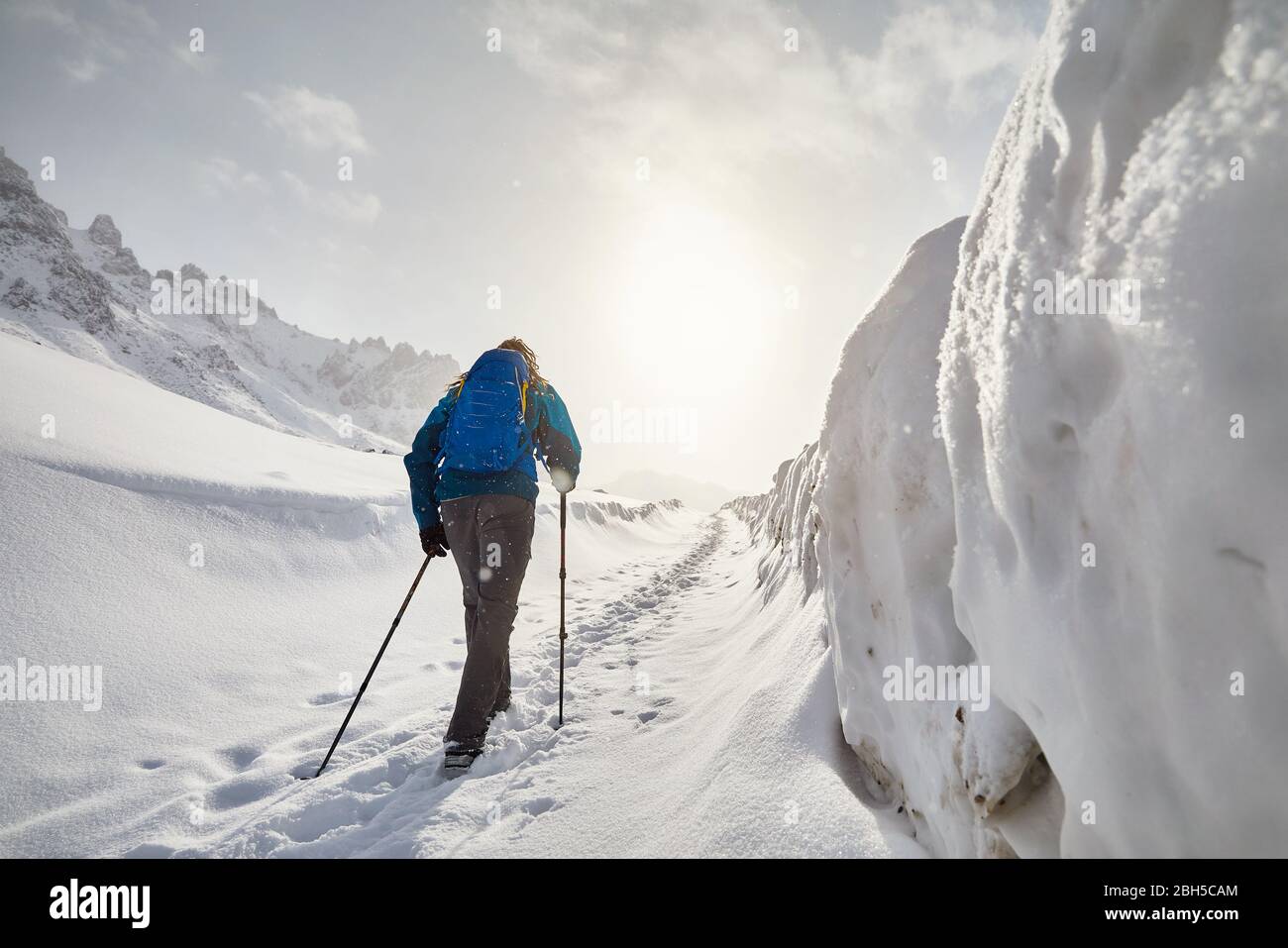 Tourist with blue backpack walking on the snow road in the mountains at snowfall Stock Photo