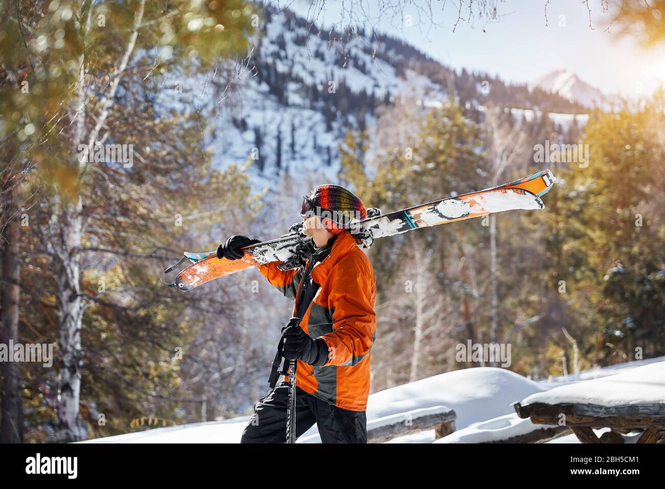 Man in orange jacket holding ski and looking at snow mountain at sunny day Stock Photo