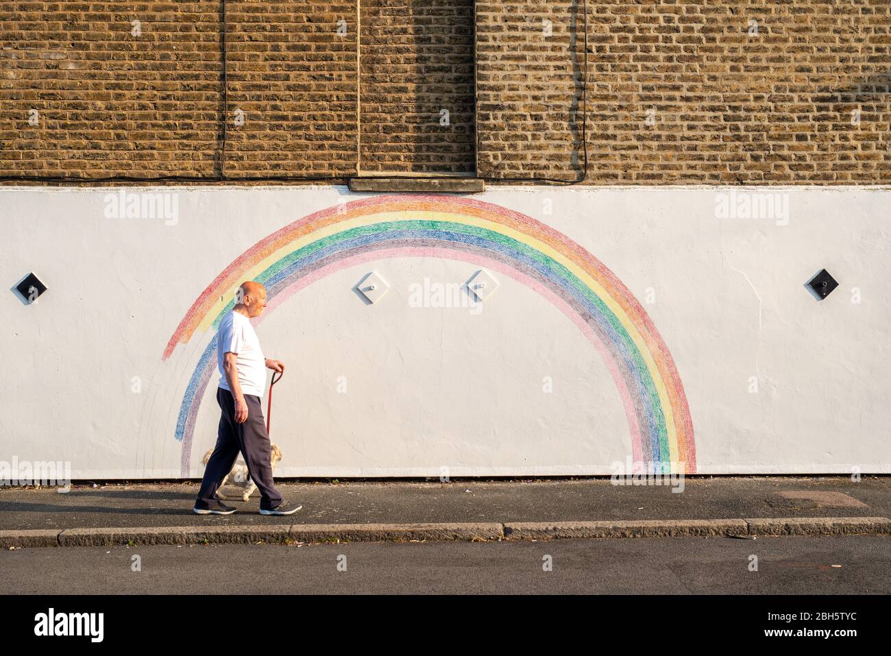 Camberwell, London, UK. 24rd April 2020. A man walks his dog past a mural of a rainbow in the early morning. Since the Covid-19 pandemic, the rainbow has become a symbol of support in the UK for NHS staff and all carers. This rainbow is being painted by Louis Young to say thank you to the NHS staff who looked after his father and recently came out of hospital. Credit: Tom Leighton/Alamy Live News Stock Photo