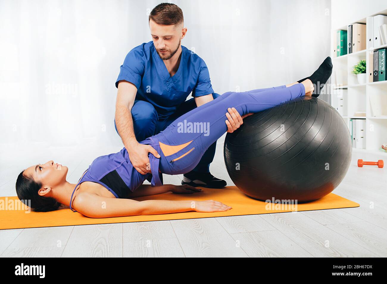 Physical therapist helping his patient doing exercise. Woman laying on mat and doing exercise with pilates ball Stock Photo