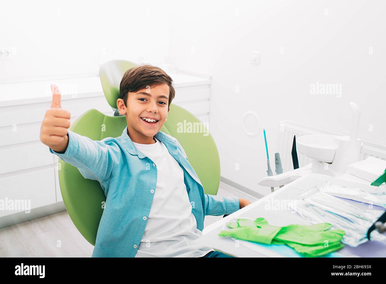 Happy little boy showing thumbs up . He sitting at dental chair, after teeth treatment. Yes, my teeth is healthy Stock Photo