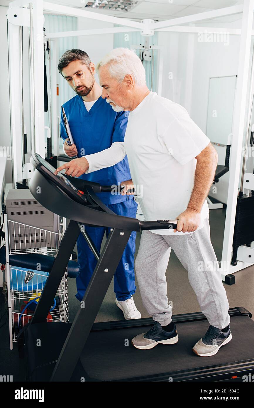 Elderly man is recovering on a treadmill. His physiotherapist helps him recover his body after physical injury. Stock Photo