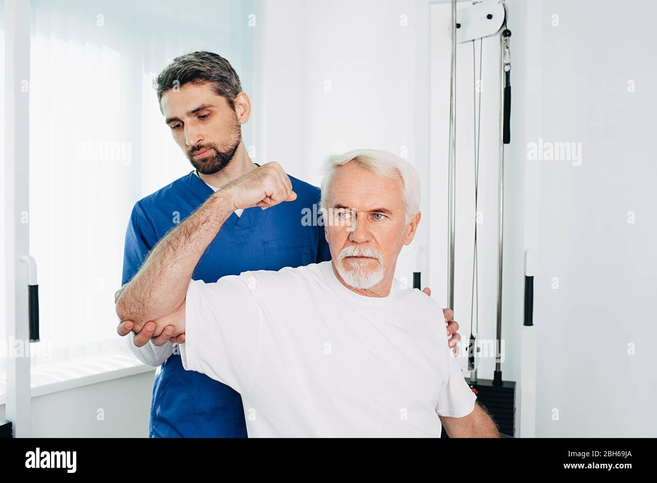 Male physical therapist holding senior patient while he stretches his cubit. Physio rehab at wellness center Stock Photo