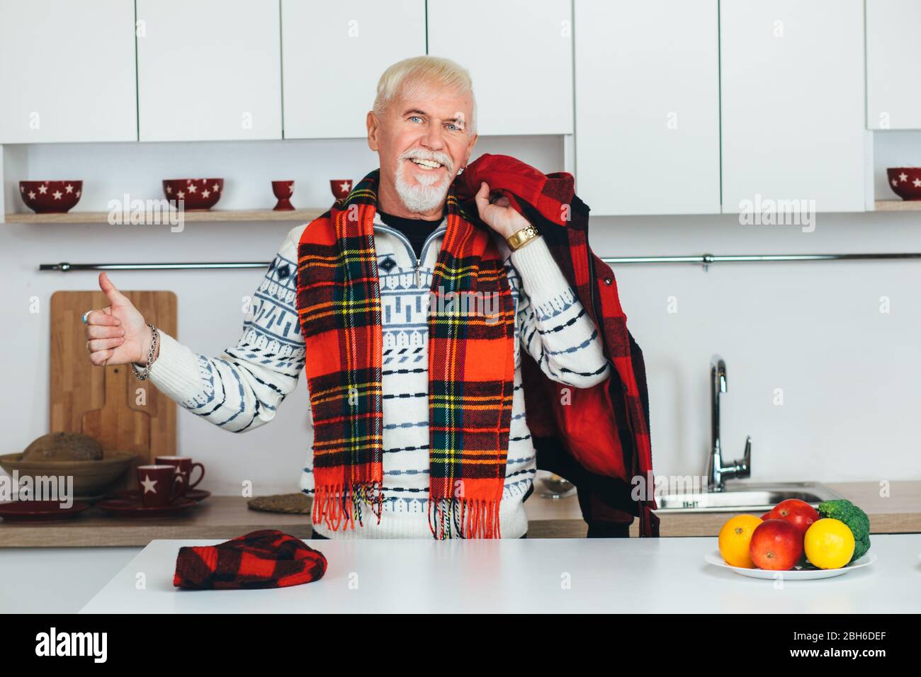 elderly man standing in outerwear, checkered scarf at home in the kitchen, looking at camera smiling and showing thumbs up Stock Photo