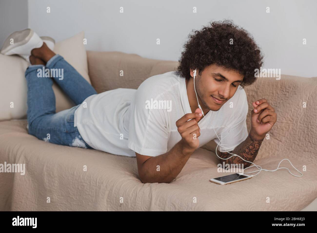 Curly haired stylish man eyes closed lying on the couch at home, enjoy the music. Stock Photo