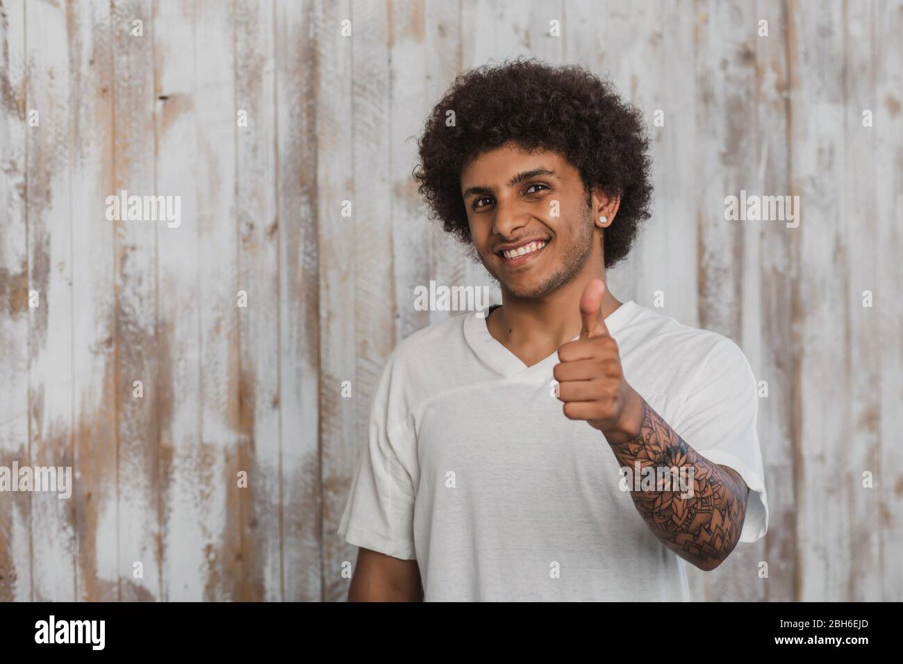 Positive curly haired man with beautiful tattoo showing thumbs up. While standing against old wooden background Stock Photo