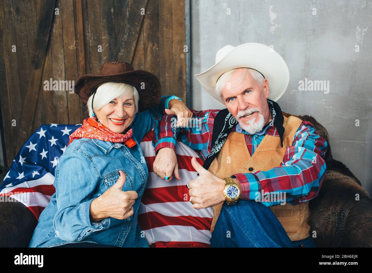 Elderly couple dressed in cowboy hats, showing thumbs up, smiling and looking at camera. Behind, on the couch lies the American flag, the celebration Stock Photo