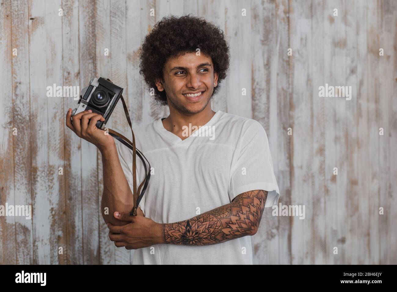 Portrait attractive and mulatto man with curly hair,holding a retro camera in his hand and smiling, behind him the old wooden background Stock Photo