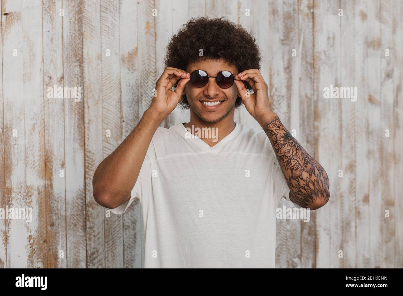 Choose a fashionable summer accessory! Portrait man with stylish hairstyle and perfect smile adjusting his sunglasses. While standing against old wood Stock Photo