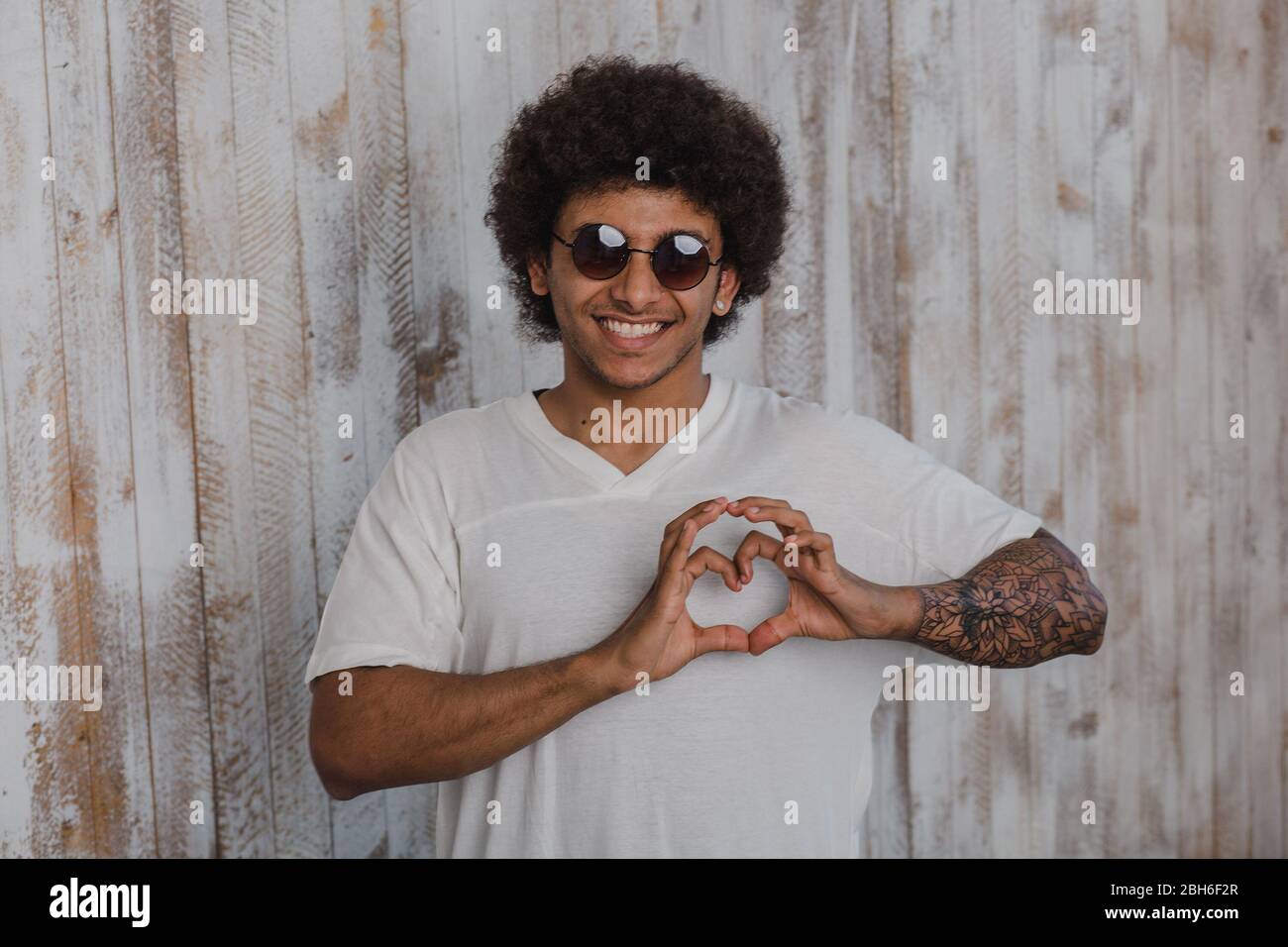 With love. Positive and curly mulatto man, wearing sunglasses, folded his hands in a heart shape, while standing against old wooden wall Stock Photo