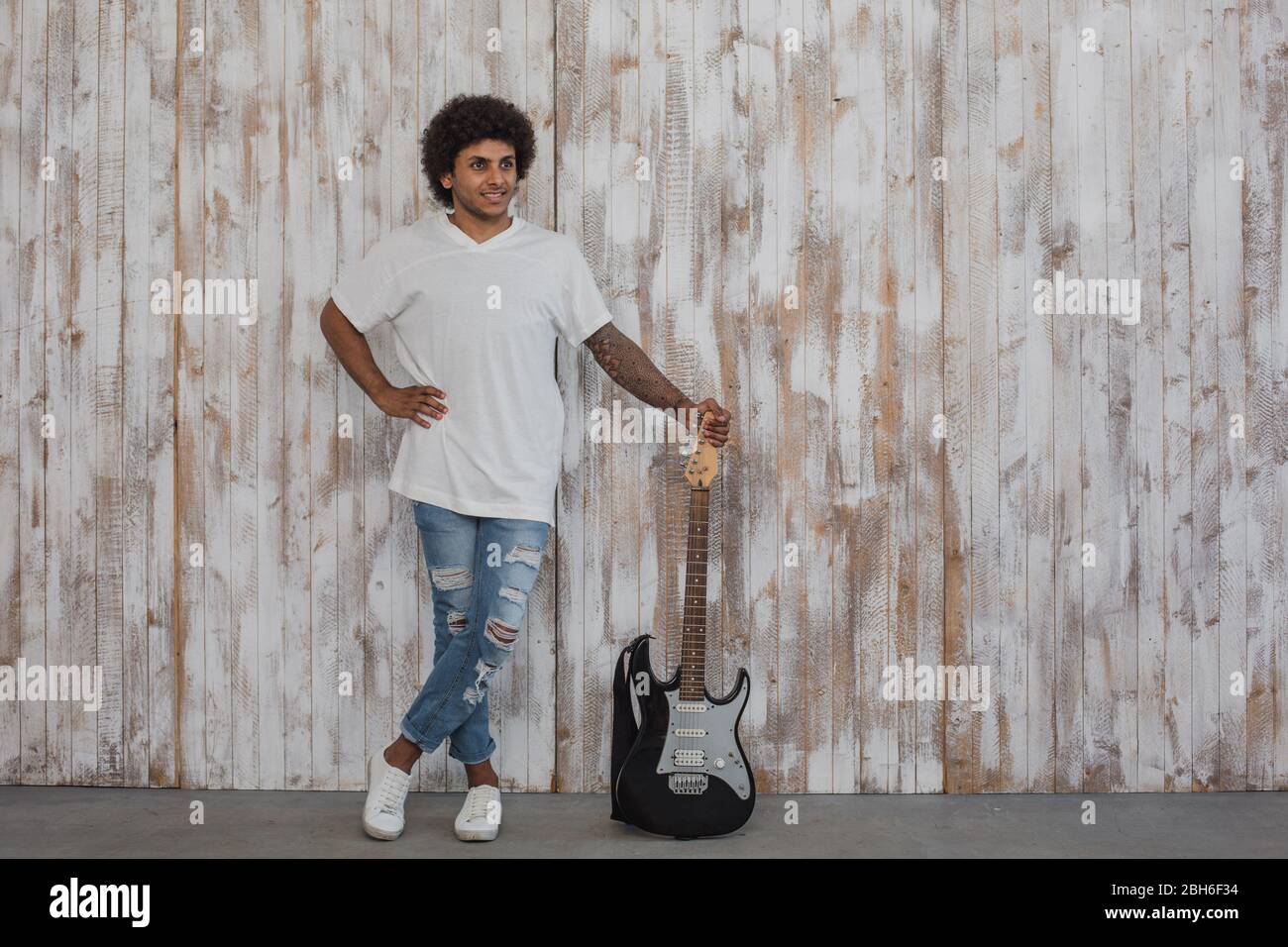 Creative person. Mulatto man musician, with curly hair stands near the bass guitar, against retro wooden wall Stock Photo