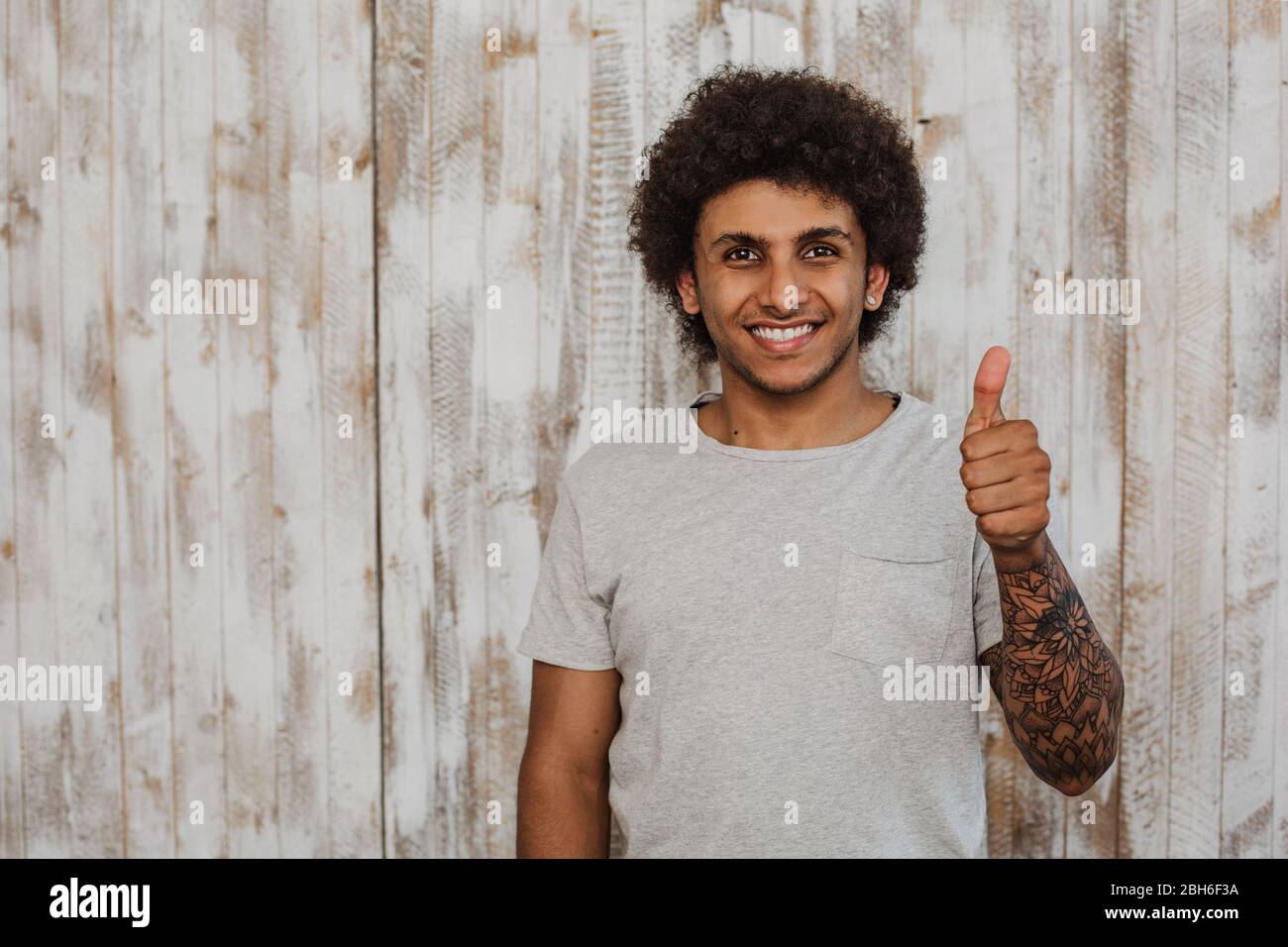 Do not worry be happy! Portrait cheerful curly haired man with perfect smile showing thumbs up. While standing against old wooden background Stock Photo