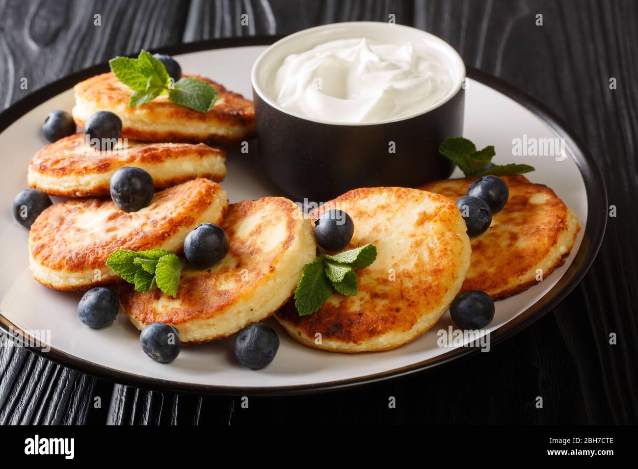 Recipe for delicious cottage cheese syrniki served with blueberries, mint and sour cream close-up in a plate on the table. horizontal Stock Photo