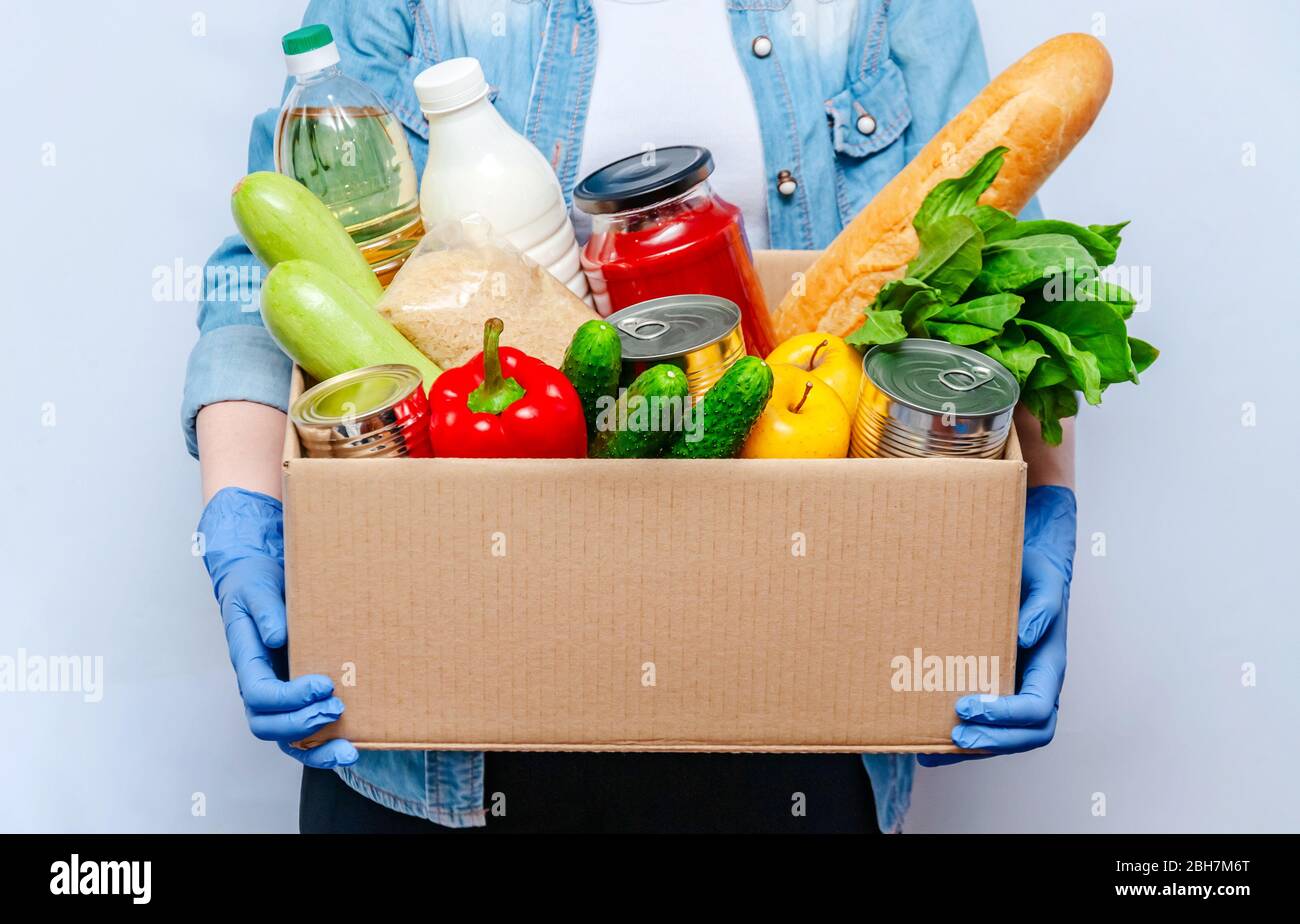 Woman in Gloves Holding Donation Box Food Supplies for People in Isolation on Yellow Background. Essential Goods: Oil, Canned Food, Cereals, Milk, Veg Stock Photo
