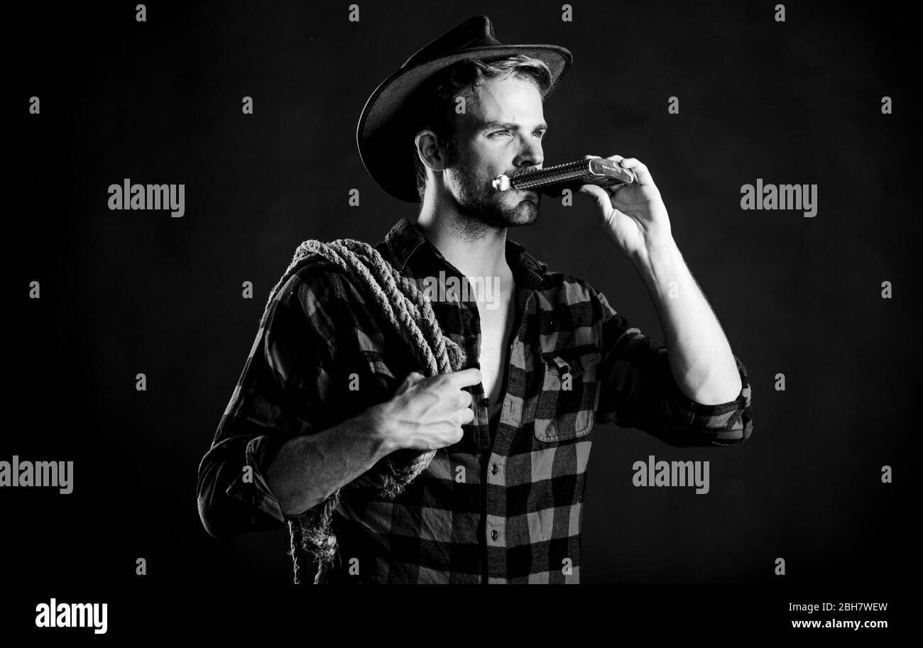 Sheriff concept. Brutal cowboy drinking alcohol. Western culture. Western life. Man wearing hat hold rope and flask. Lasso tool of American cowboy. Man handsome unshaven cowboy black background. Stock Photo