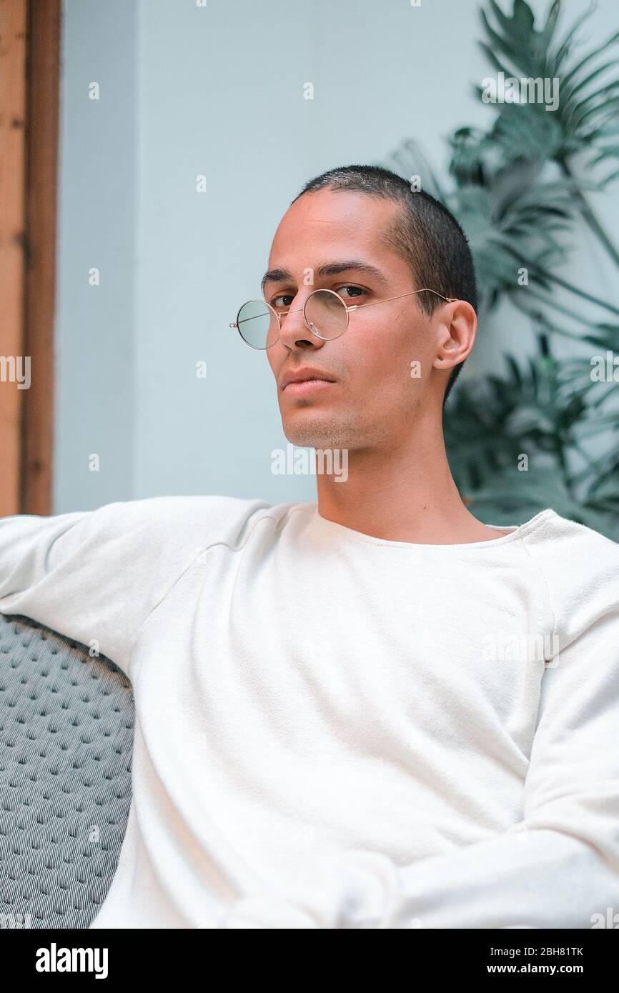 Exotic caucasian young man with round transparent sunglasses looking to the camera while sitting on a sofa in front of a tropical plant Stock Photo