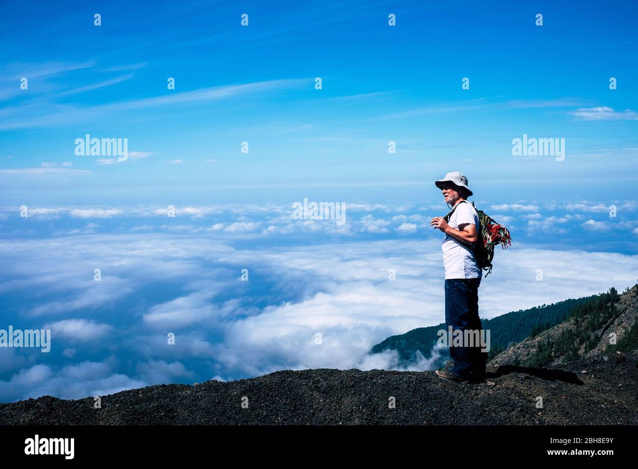 Lonely retired adult white hair man standing during a trekking travel adventure with his backpack and loooking the beautiful landscape of sea of clouds in front of him. amazing nature Stock Photo
