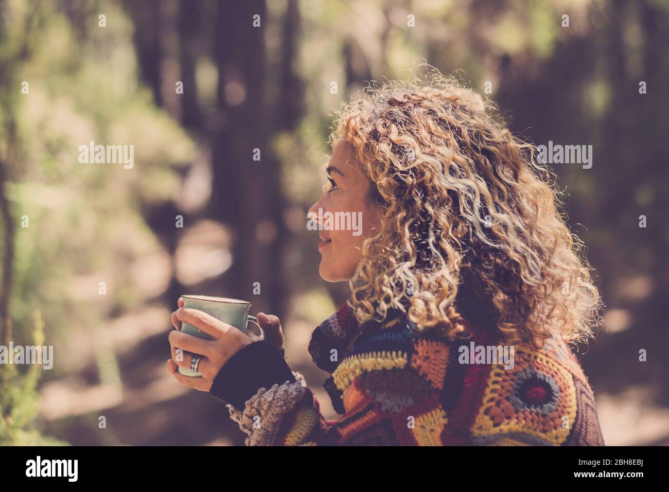 Beautiful woman in autumn forest enjoying the feeling with the nature with a warm sweater - lady sits with trees in nackground in a forest and holds a cup with a hot drink in her hands. Girl travel and wanderlust millennial concept Stock Photo
