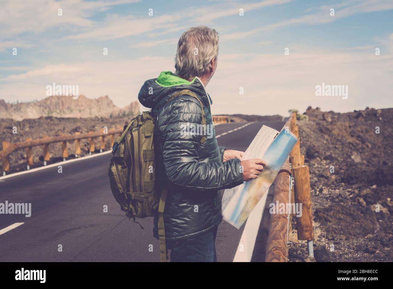 portrait of adventure senior man with map and extreme explorer gear on mountain with long straight road in front of him for a long walk. wanderlust and travel concept image. rocks and blue sky scenic place Stock Photo
