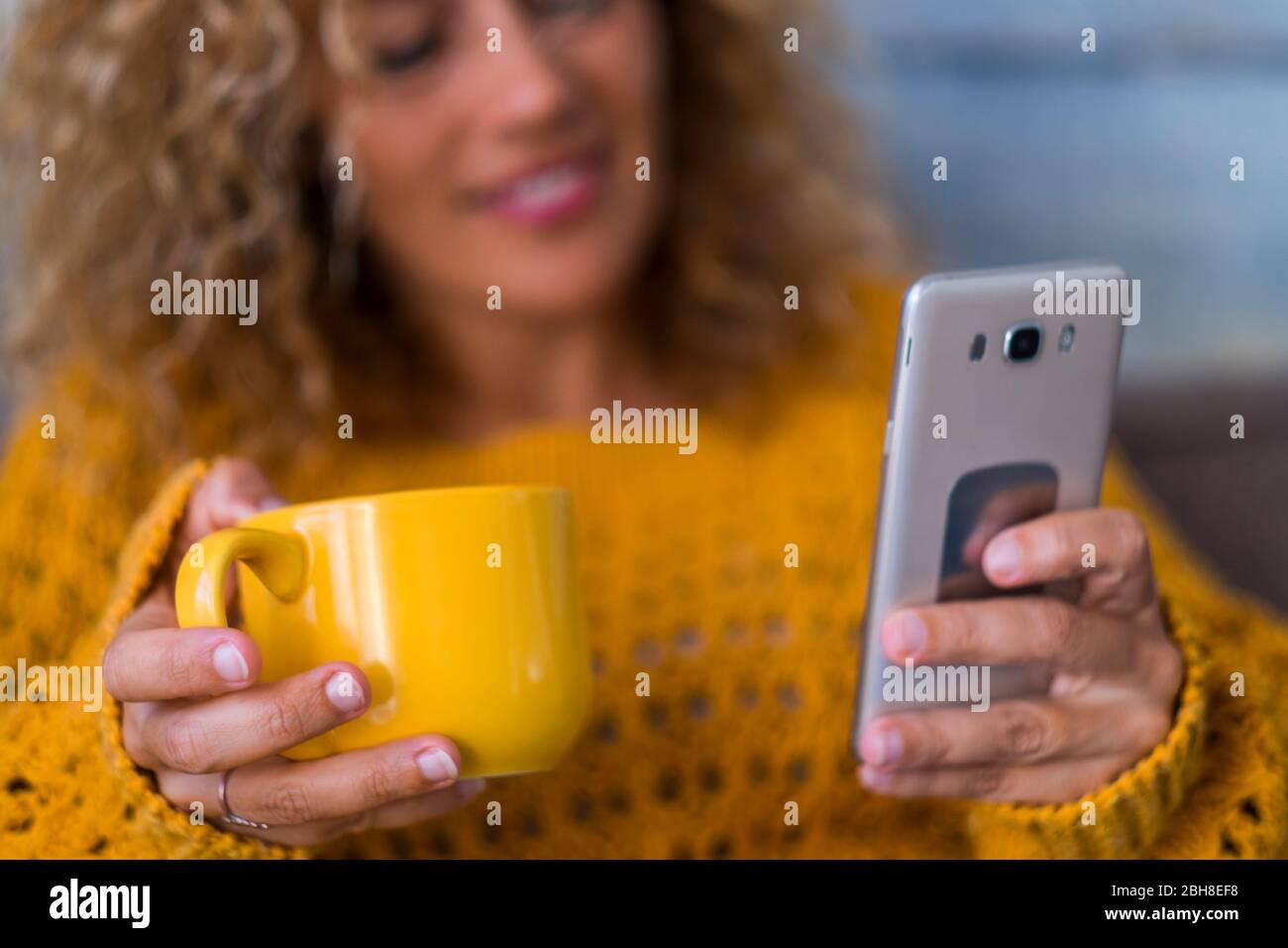 Autumn and technology mobile phone cconcept - middle age modern woman drink tea or coffee at home while chek the cellular for internete messages od news from social media - focus on mug Stock Photo