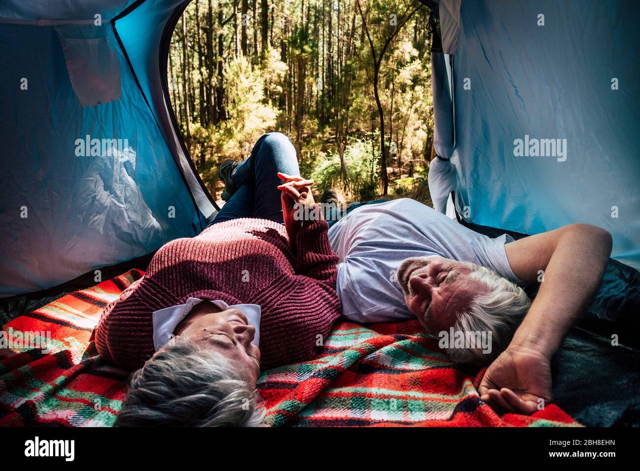 adult senior couple rest lay down inside a tent in free wild camping in the forest for alternative travel and lifestyle. Love forever together concept for man and woman taking hands andd look eachother - outdoor nature activity Stock Photo