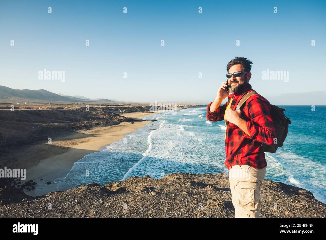Standing caucasian man calling at the phone on a isolate cliff with ocean waves view - backpacker alternative lifestyle vacation for people love nature and outdoors - technology and adventure concept Stock Photo