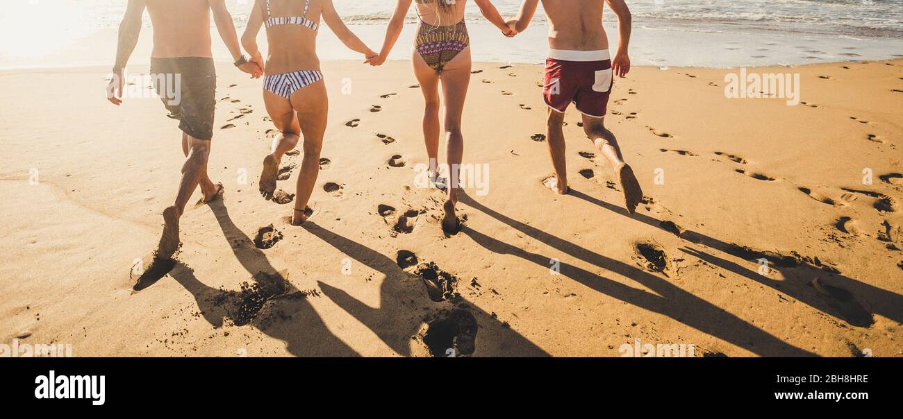 Back view people in summer holiday vacation running on the sand at the beach to the water sea all together in friendship holding hands - bikini group men and women have fun in the sunset with shadows Stock Photo