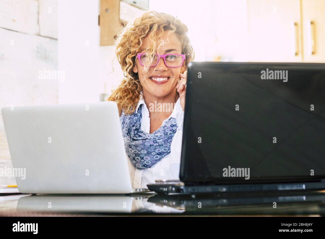Beautiful blonde curly adul twoman wotk at the office with two laptop notebook at the same time - people at work concept - businesswoman using technology to work online at home Stock Photo