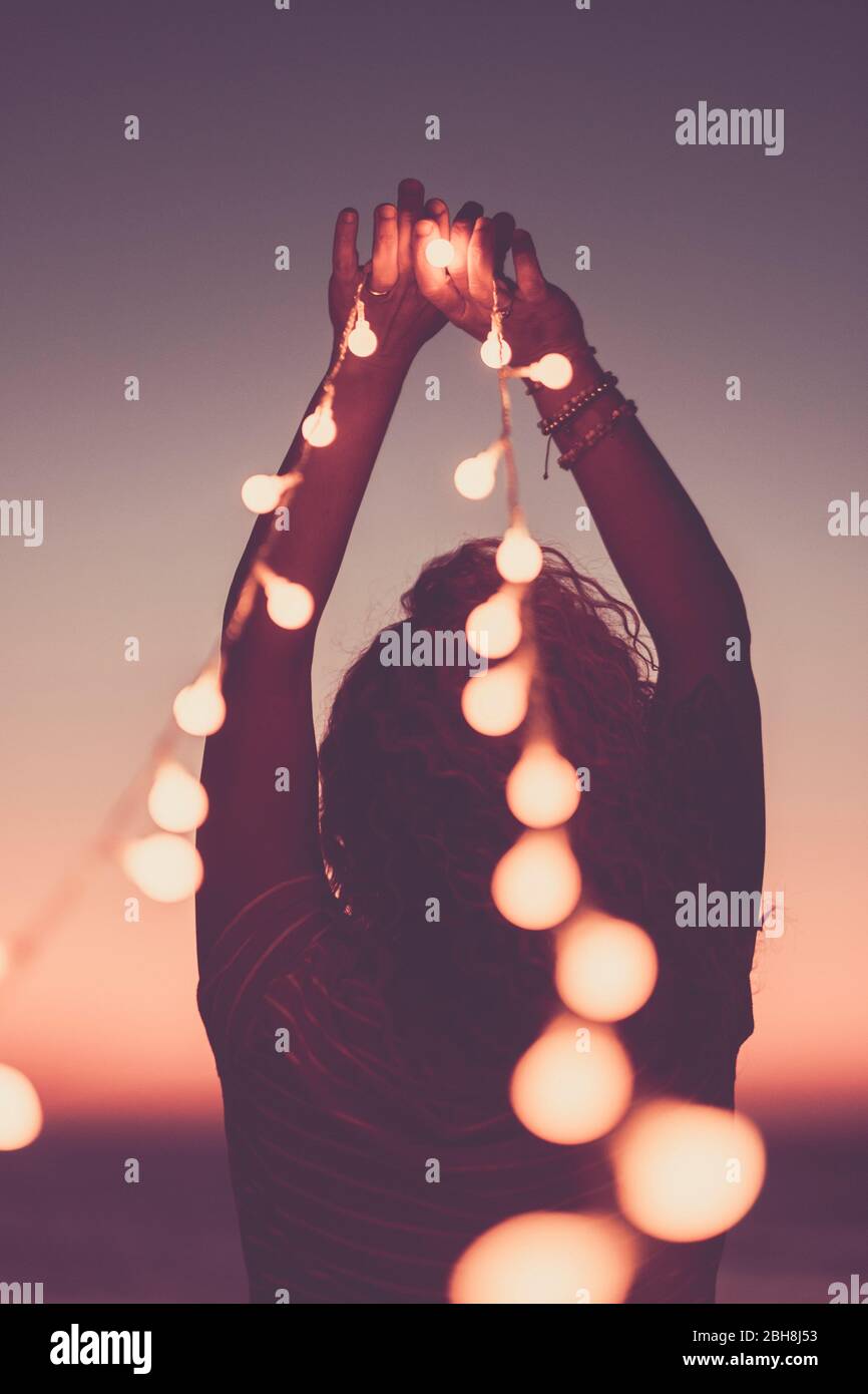 Motivational inspirational concept image with girl from back holding yellow bulb lights to the sky - pink coloured style filter - hope and feeling - lifestyle for happy people - sunset in background Stock Photo