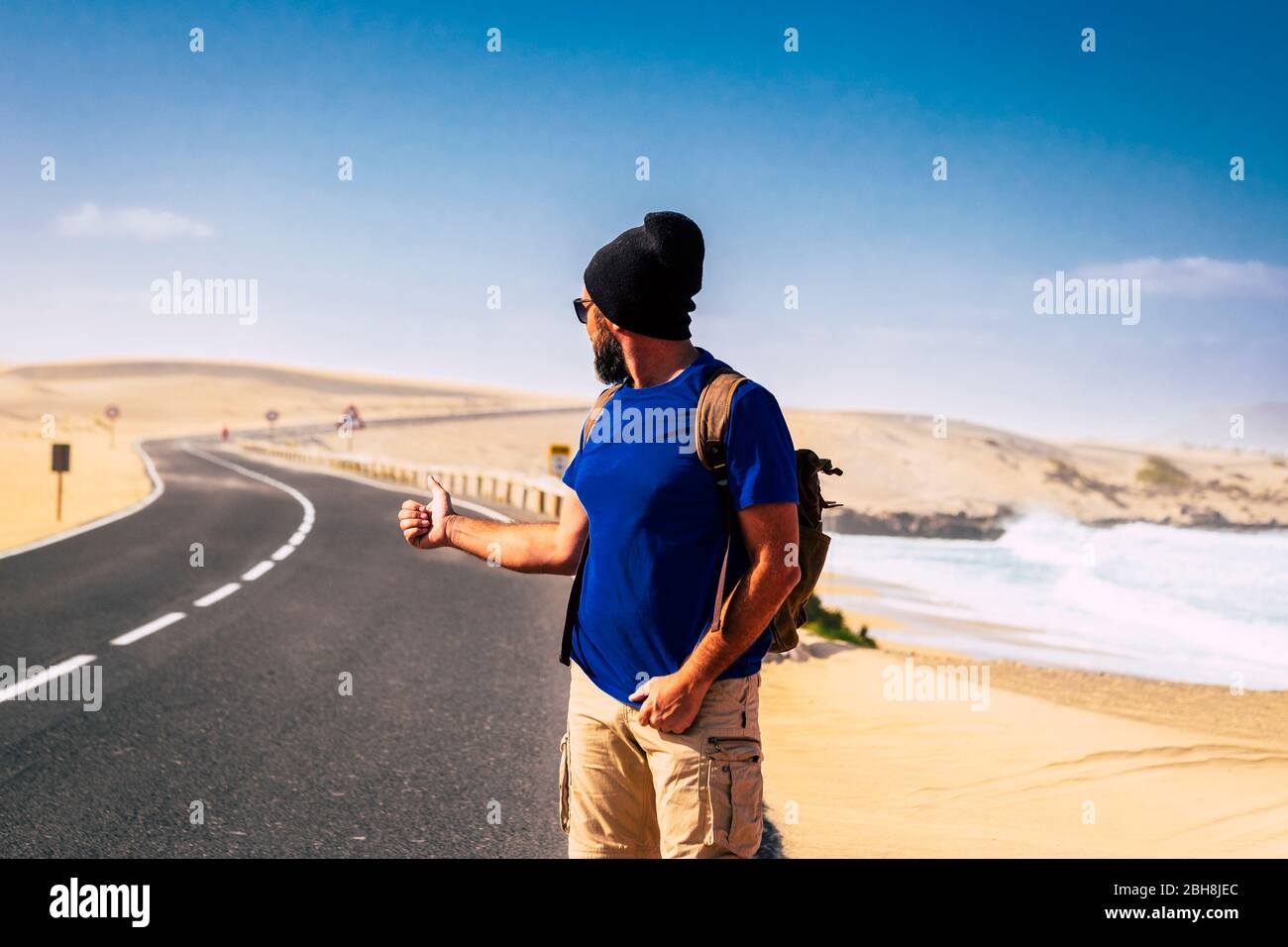 Hitcher alternative travel people concept with man with a backpack waiting for a car to share the trip - lonely travelers with long road and desert and sandy beach in background - holiday wild adventure Stock Photo