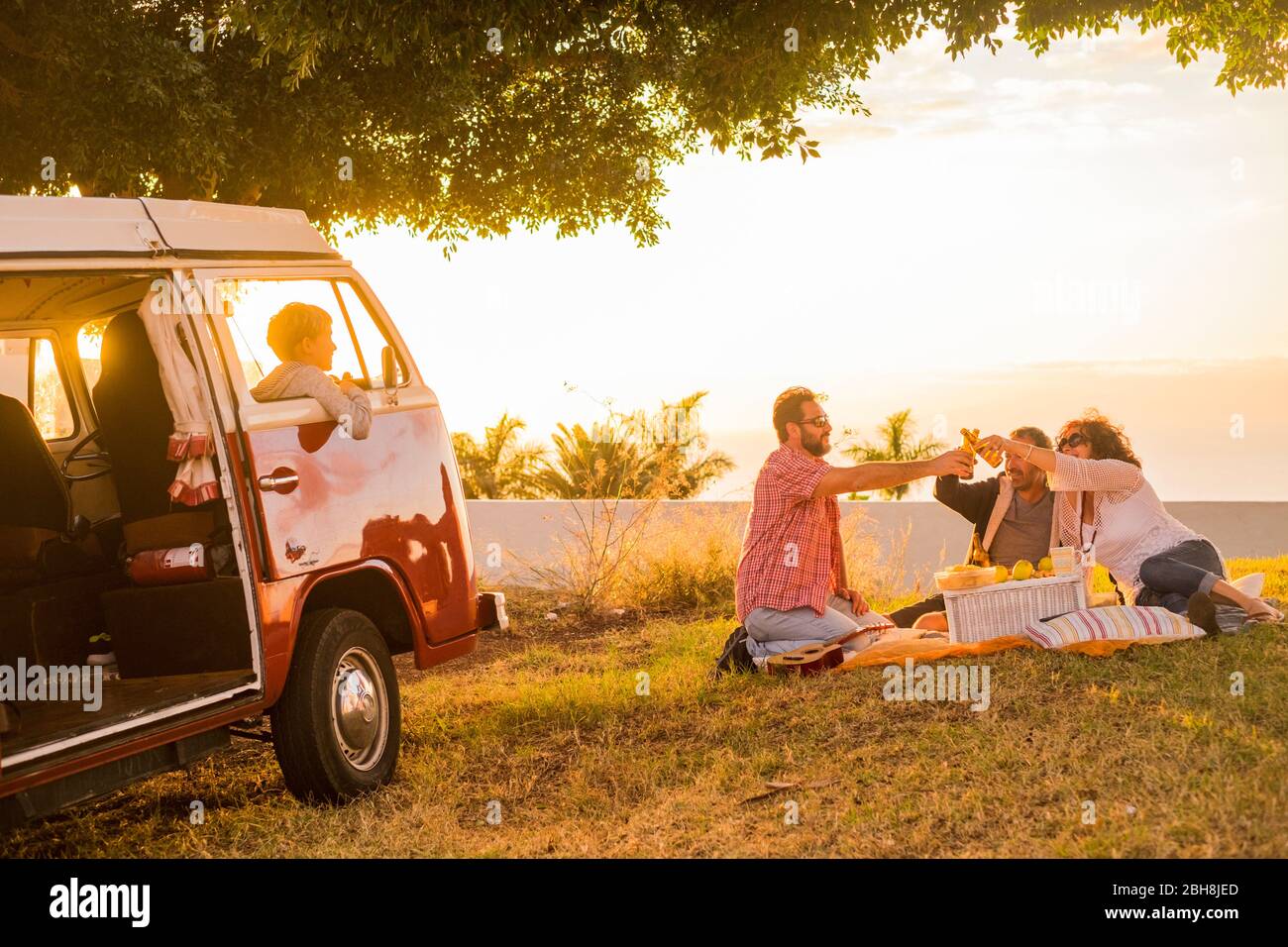 Family and friends all together in picnic leisure activity on a meadow with a red old vintage van parked and son children inside loooking them clinking with some beers during a coloured golden sunset Stock Photo