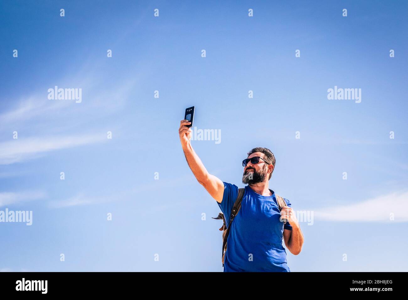 People and internet technology man with beard and sunlgasses loooking for signal with a mobile phone device - people traveling with backpack for adventure concept and alternative vacation - blue sky in background Stock Photo