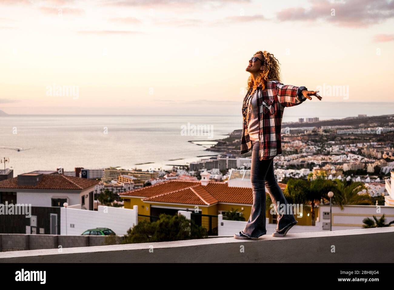 Creativity happiness freedom and craziness concept with young beautiful curly blonde woman walking in balance on a wall with city and coastline down in the background - cheerful people enjoy Stock Photo