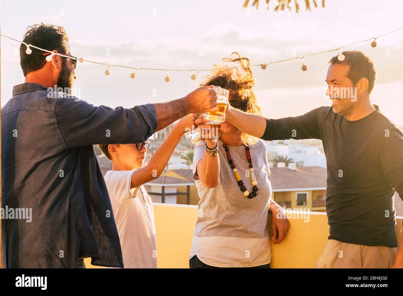 Group of friends toasting and clinking glasses with beer and orange juice together during the sunset on the roof terrace with city view - mixed ages and generations having fun celebrating Stock Photo