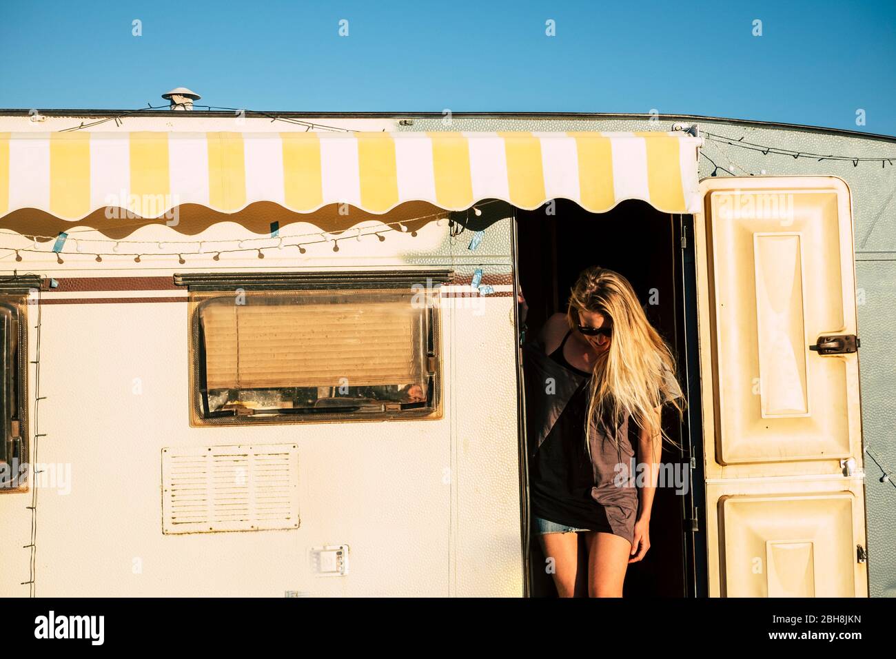 Old style american concept with beautiful blonde young girl on the door of a vintage old caravan - diversity and alternative travel style and vacation holiday adventure - freedom and tiny house Stock Photo