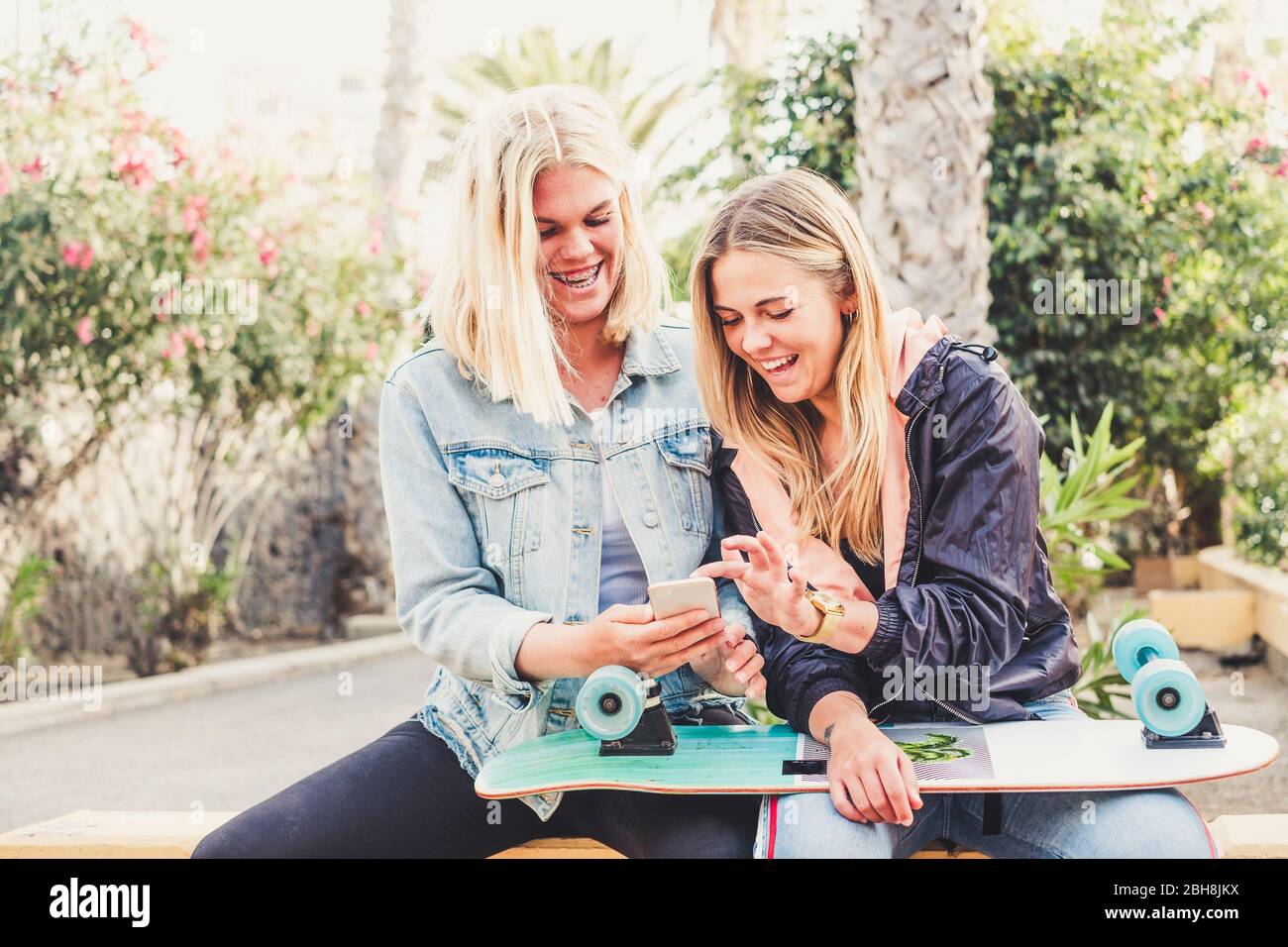 Couple of friends having fun using technology and smartphone witting in a park - hipster skateboard and millennial people in friendshp together - laugh and smile blondes girls Stock Photo