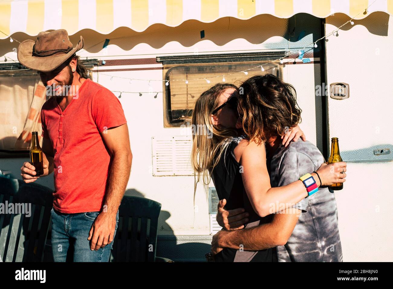Couple of young people kissing with love with a friend walking in the back and smiling - youthful generations like travel and enjoy the outdoor adventure - old caravan in background Stock Photo