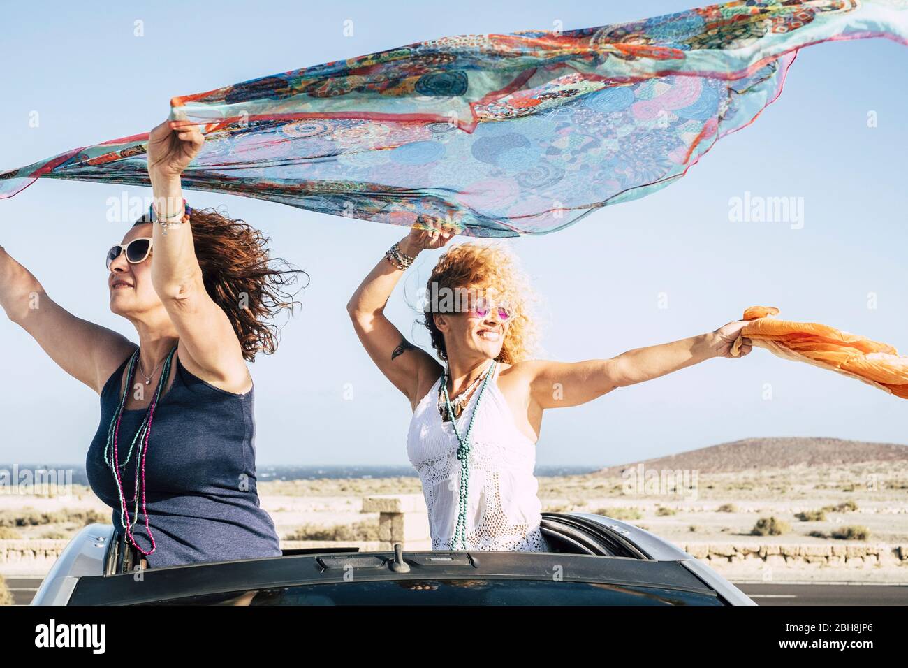 Couple of women have fun and play with the wind standing out of the top of a convertible car and playing with the wind with coloured pareos - cheerful people traveling and enjoying the outdoor on vacation Stock Photo