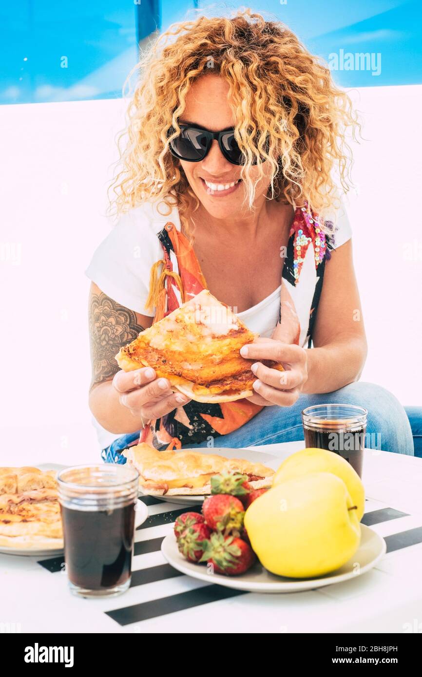 Beautiful cheerful curly long blonde hair woman eating italian pizza outdoor in restaurant or home terrace - trendy young female with tatoo and casual coloured style Stock Photo