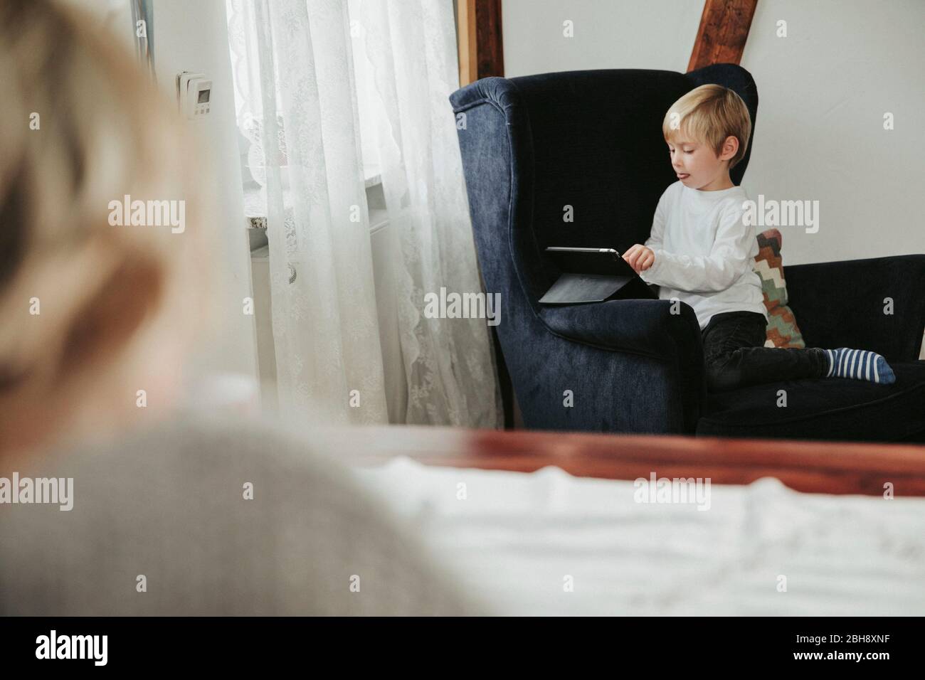 kleiner Junge mit Tablet Computer im Sessel, Mutter unscharf im Vordergrund Stock Photo