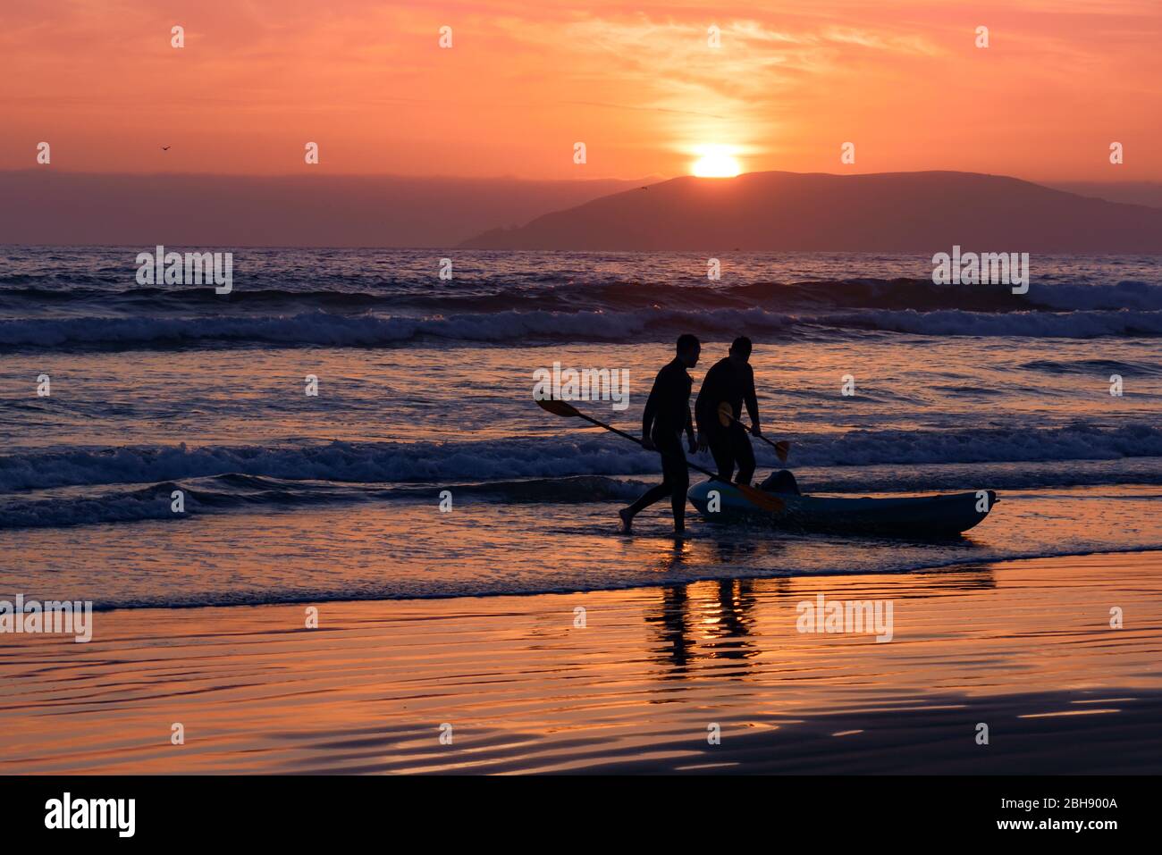 Silhouette von 2 Paddlern mit Boot am Strand bei Sonnenuntergang, im Hintergrund die Silhouette von Hügeln, die die Bucht um Pismo Beach umranden Stock Photo