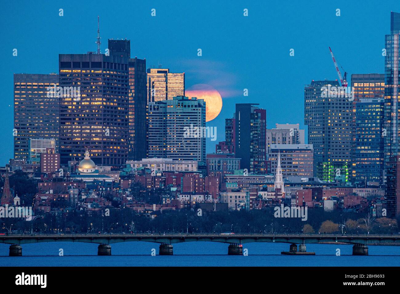 Moon rising beyond city peaking behind buildings at blue hour. Stock Photo